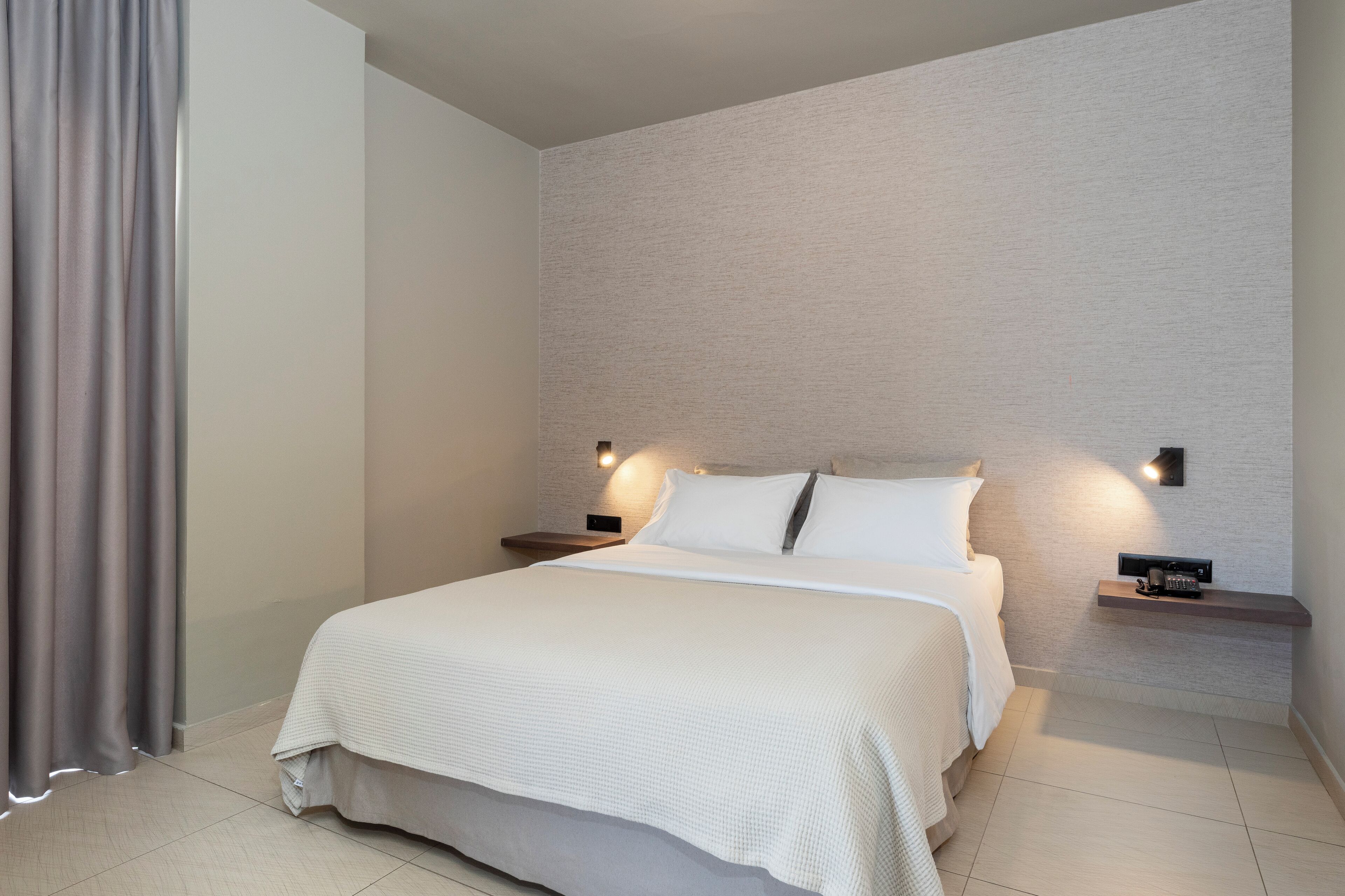 Una cama bien hecha con edredón blanco y almohadas, dos mesitas de noche con lámparas y una pared de color claro con textura.