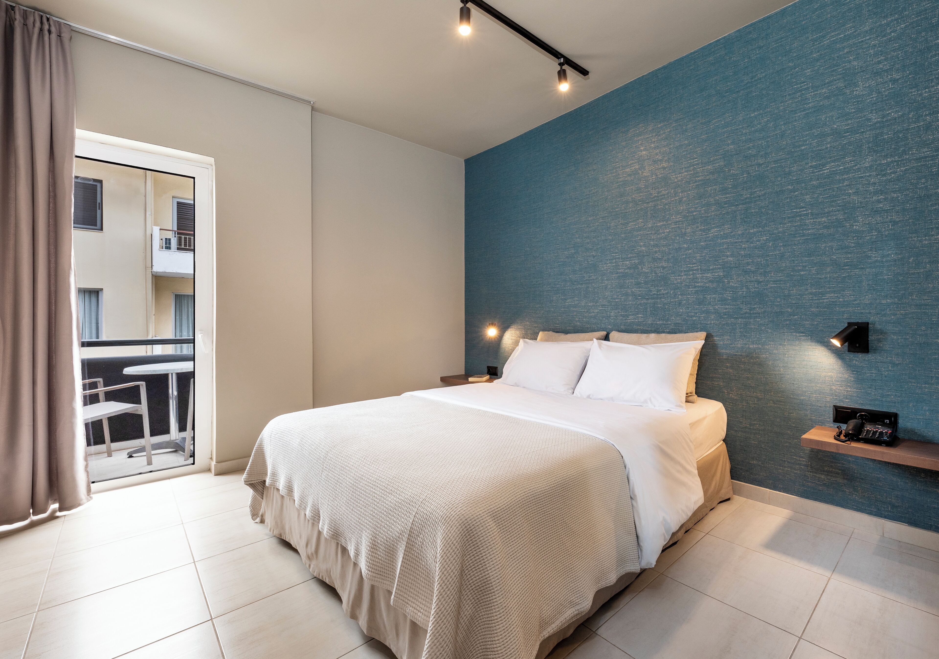 A modern bedroom with a large bed, a balcony with a table and chairs, and a blue accent wall.