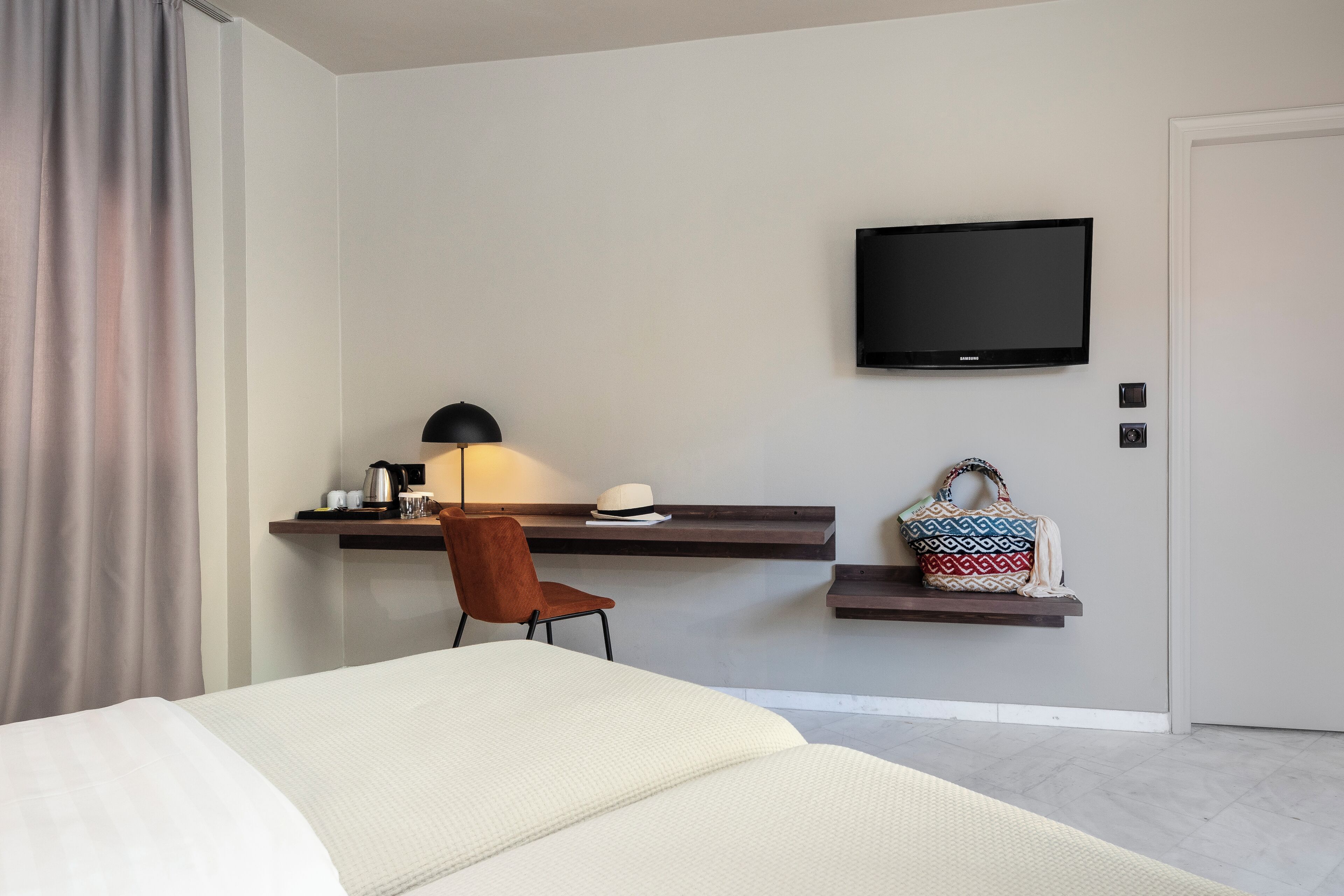 A modern hotel room with a bed, a desk with a lamp, a TV, and a handbag on a shelf.