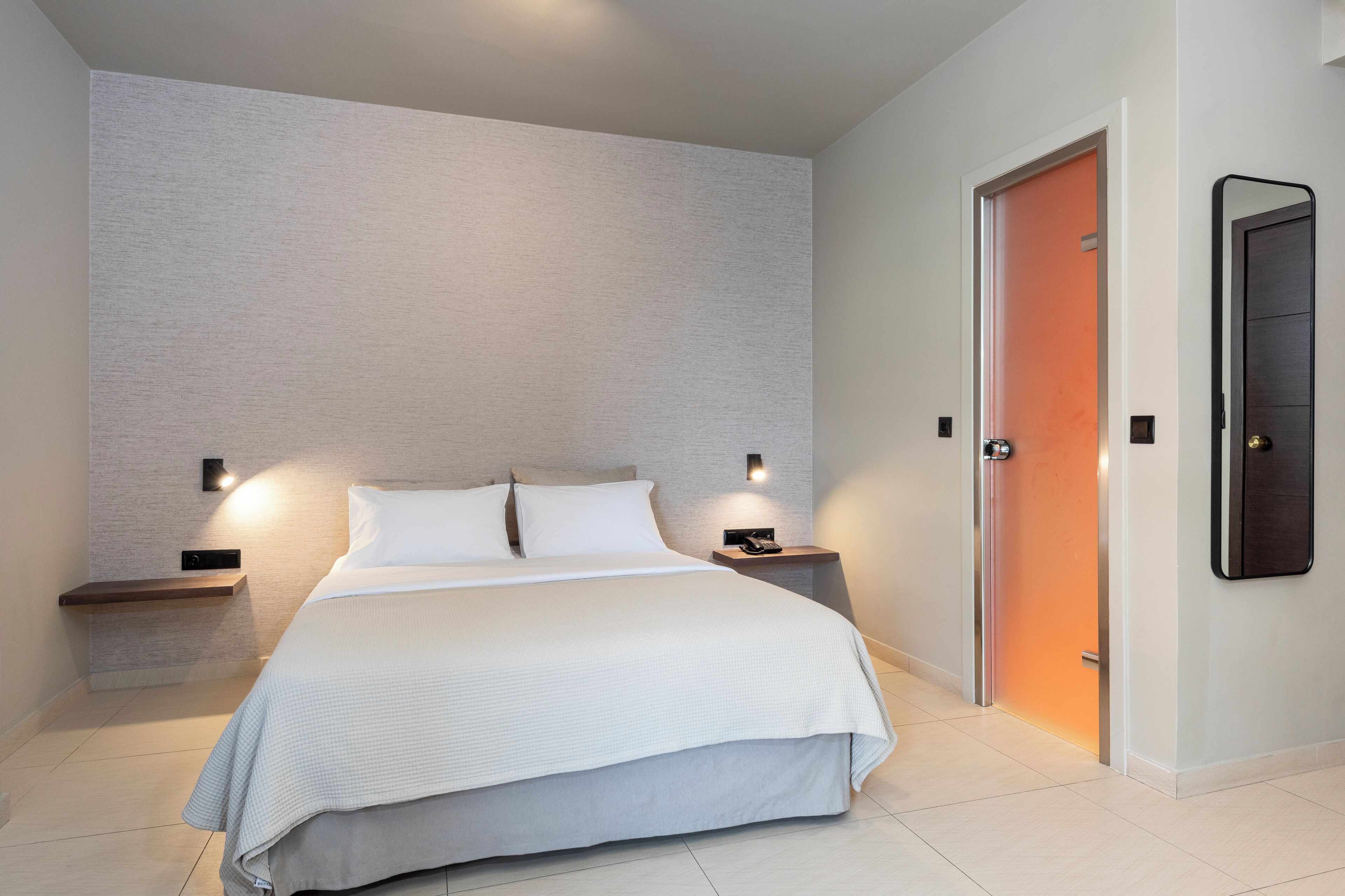 A modern hotel room with a large bed, bedside tables, a wall-mounted TV, and a door with an orange frame.