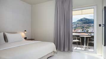 Un dormitorio con cama, mesita de noche, luz empotrada en la pared, balcón con mesa y sillas, y vistas a un pueblo y una colina.