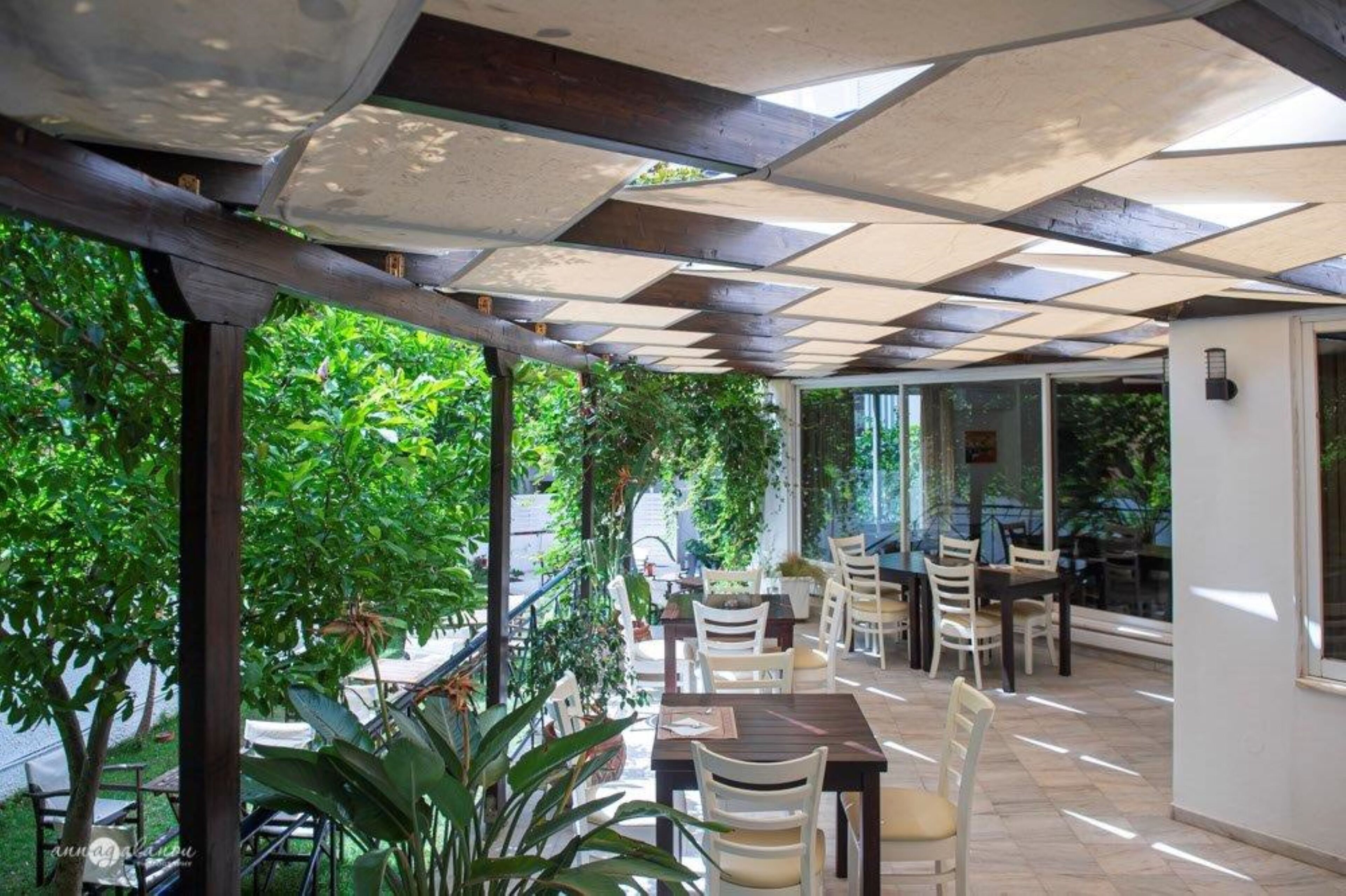 Un patio exterior con mesas y sillas, rodeado de follaje y un techo a cuadros.