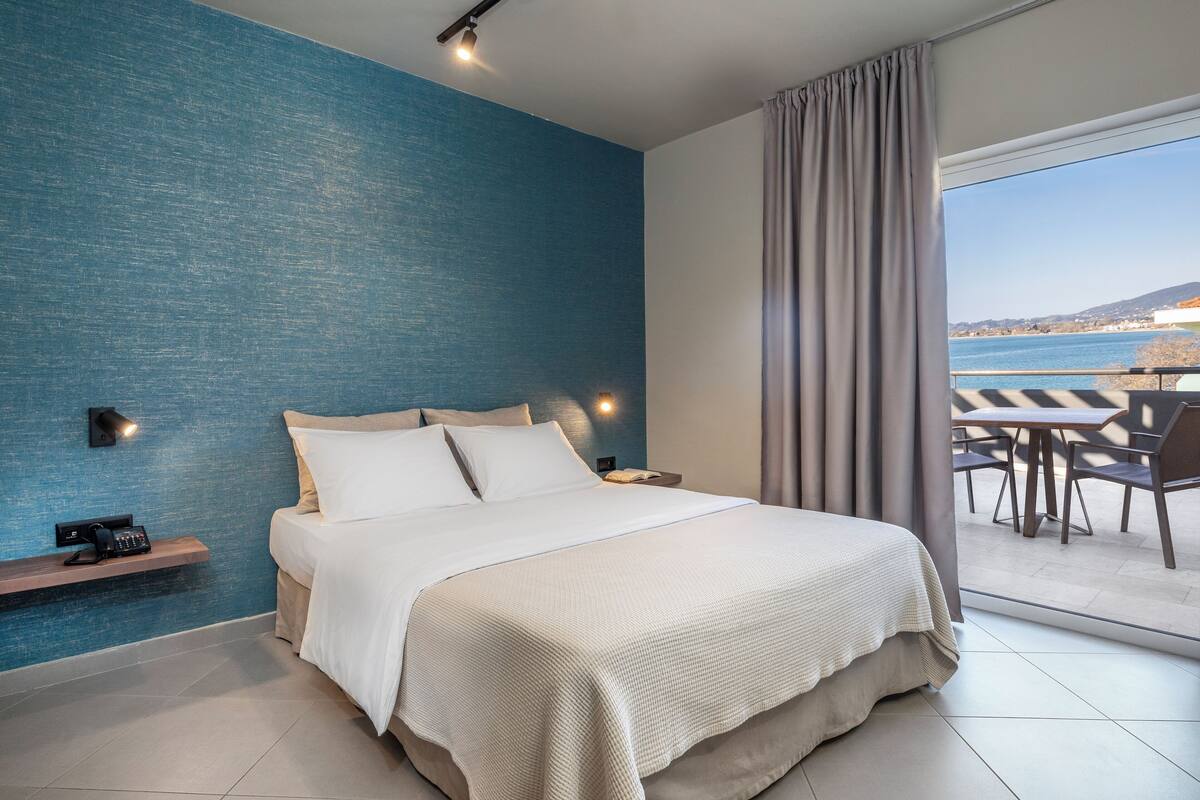 A modern hotel room with a large bed, a bedside table with a phone, a blue textured wall, and a view of the sea through a balcony.