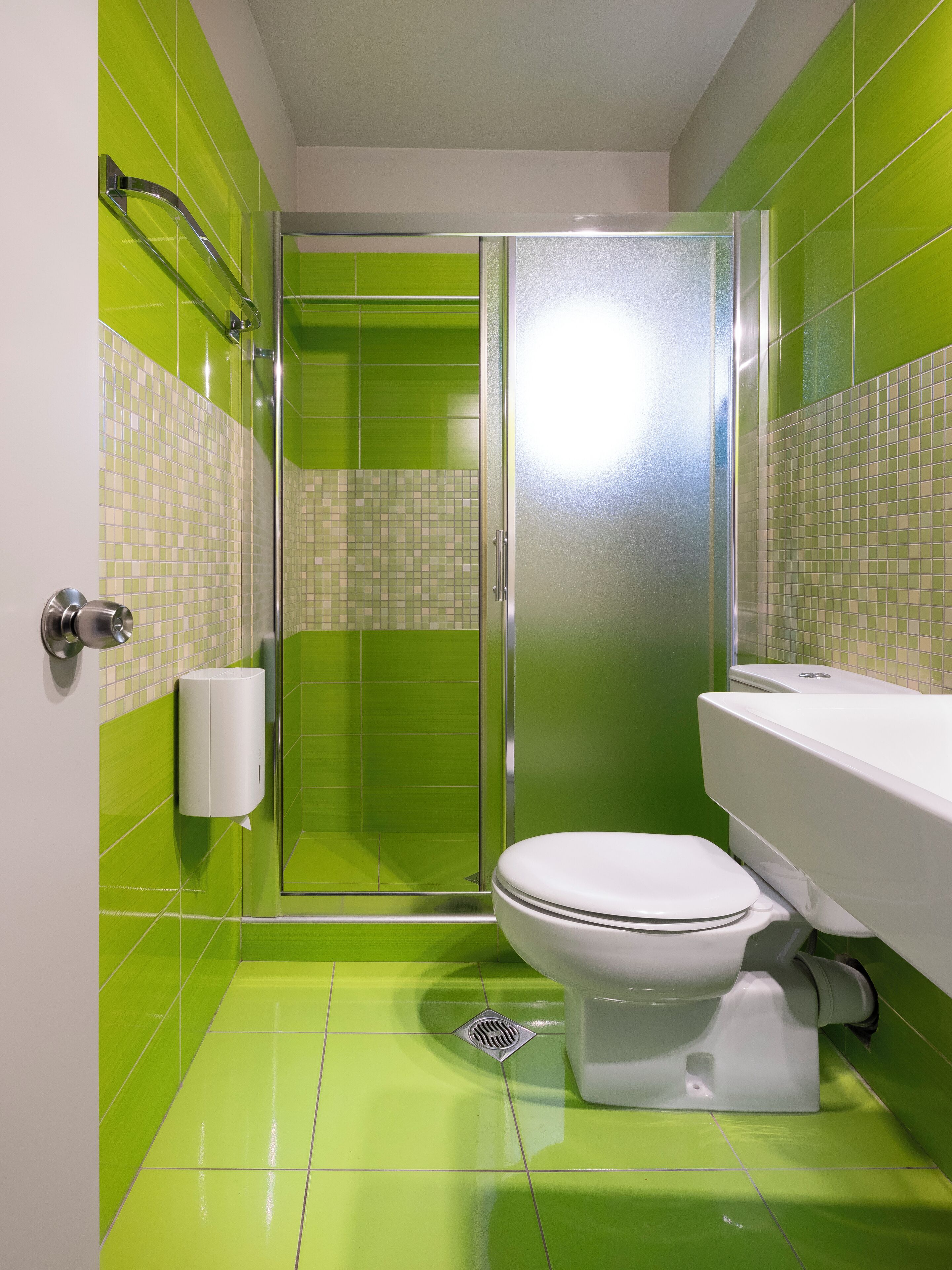 A bathroom with green tiles, a white toilet, a glass shower enclosure, and a white sink.