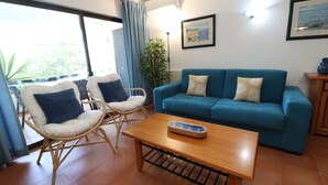 A living room with a blue sofa, two wicker chairs, a wooden coffee table, and a large window with curtains. - 2 Bedroom Apartment with Swimming pool at São Rafael Beach - Albufeira (Albufeira)