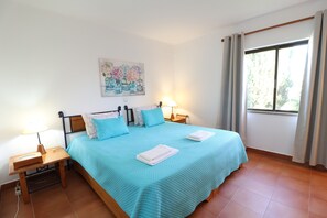 A bedroom with a bed, two bedside tables, a nightstand, a wall-mounted painting, and a window with curtains.
