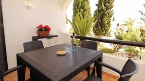 A dark wicker table with four chairs, a bottle of water, a plate of food, and a potted plant with red flowers on a balcony. - 2 Bedroom Apartment with Swimming pool at São Rafael Beach - Albufeira (Albufeira)