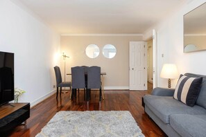 Ein Wohnzimmer mit grauem Sofa, einem Fernseher, einem Esstisch mit Stühlen und einem Teppich.