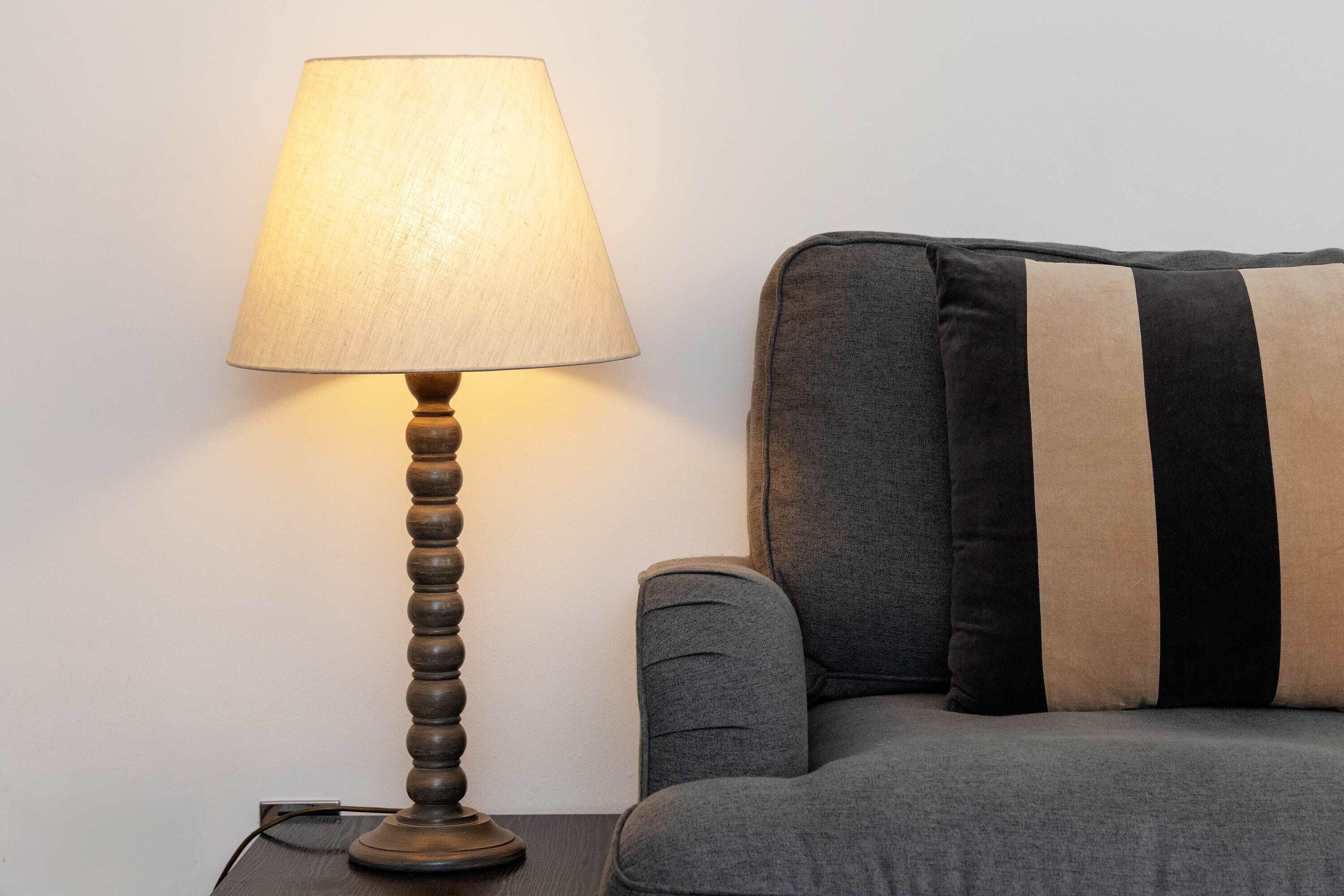 Una lámpara con pantalla color crema sobre una base de madera, ubicada junto a un sofá gris con cojines a rayas negras y color crema.