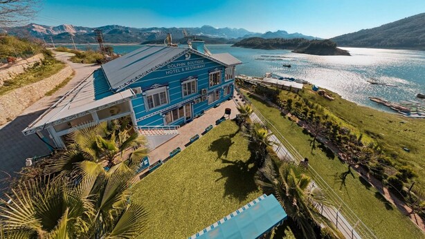 A blue building with a sign reading "DOLPHIN BOUTIQUE HOTEL & RESTAURANT" overlooking a lake with mountains in the background. - Dolphin Boutique Hotel & Restaurant (Bucak)