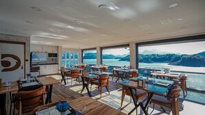 A dining area with tables and chairs, large windows with a view of a lake and mountains, and a kitchen area with cabinets and a countertop. - Dolphin Boutique Hotel & Restaurant (Bucak)