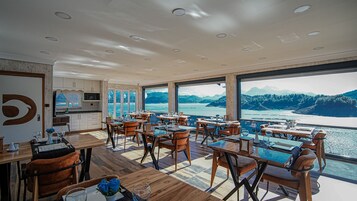 Un comedor con mesas y sillas, ventanas amplias con vistas a un lago y montañas, y una zona de cocina con armarios y encimera.
