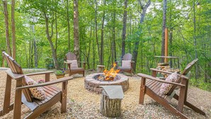Uma área externa arborizada com uma fogueira circular rodeada por cadeiras de madeira estilo Adirondack.