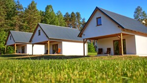A row of three white houses with grey roofs, set on a grassy lawn with trees in the background. - Źródlo Raduni (gmina Stezyca)
