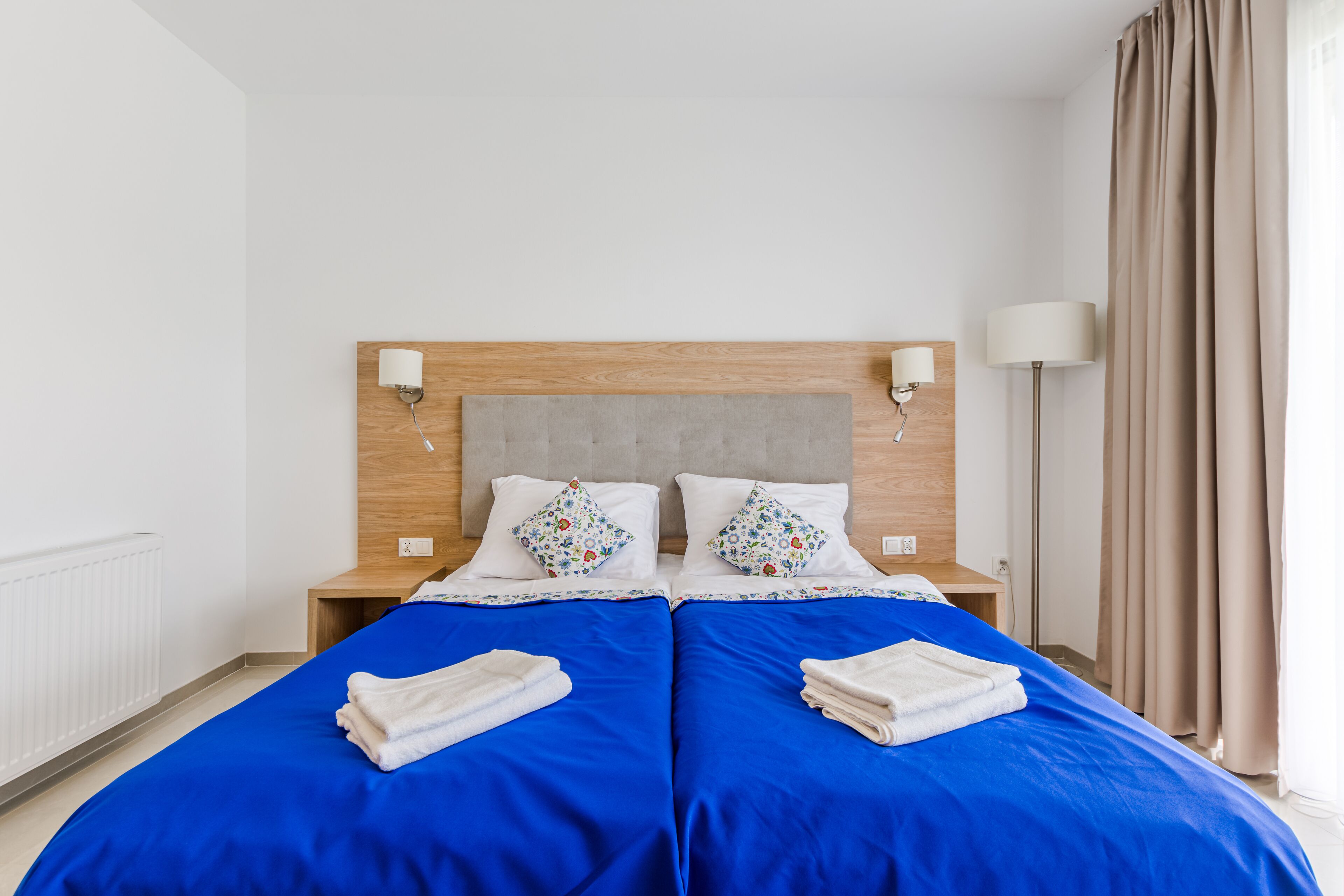A double bed with blue bedding and two pillows, a wooden headboard, bedside lamps, and a window with curtains.