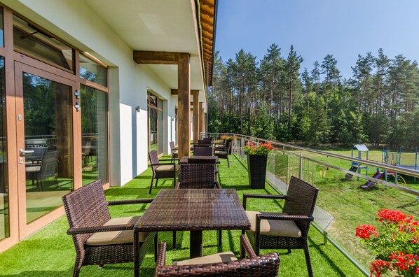 A patio with wicker furniture, a glass door entrance, and a playground in the background. - Źródlo Raduni (gmina Stezyca)