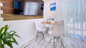 A modern room with a large TV mounted on a wooden panel, a round table with chairs, a potted plant, and a view of the outdoors through sheer curtains. - Charming Ground Level Resort Style 3 unit Condo  - unit C (Holmes Beach)