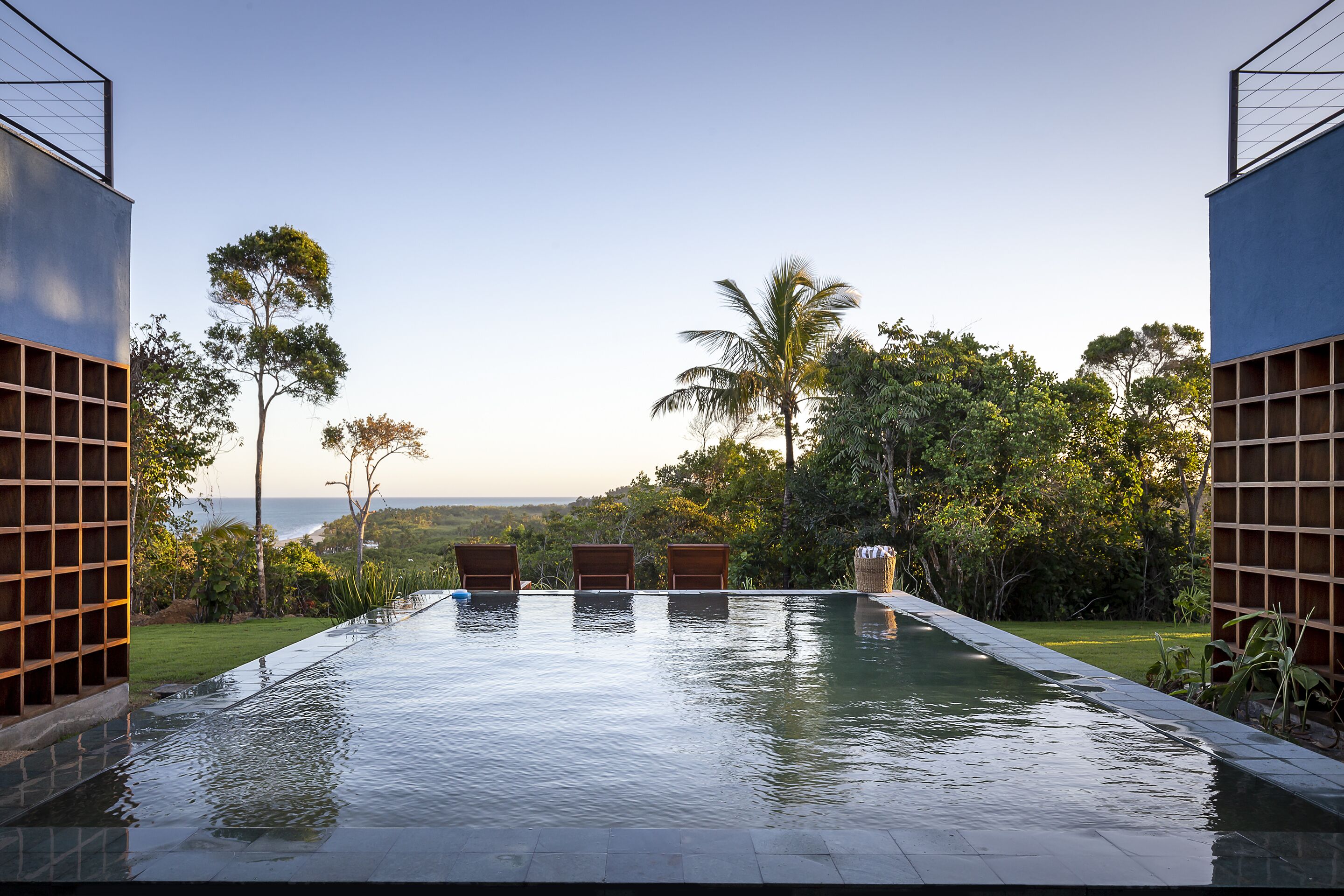 En rektangulär infinitypool med tre träloungestolar, omgiven av frodig grönska och med en fantastisk utsikt över havet.