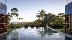 A rectangular infinity pool with three wooden loungers, surrounded by lush greenery and a clear view of the ocean. - Lux & Modern Villa w/ 5 bedrooms in Trancoso (BA)