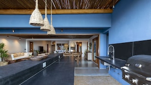 A modern kitchen with a built-in grill, a long counter, hanging light fixtures, and a dining area with a sofa and table. - Lux & Modern Villa w/ 5 bedrooms in Trancoso (BA)
