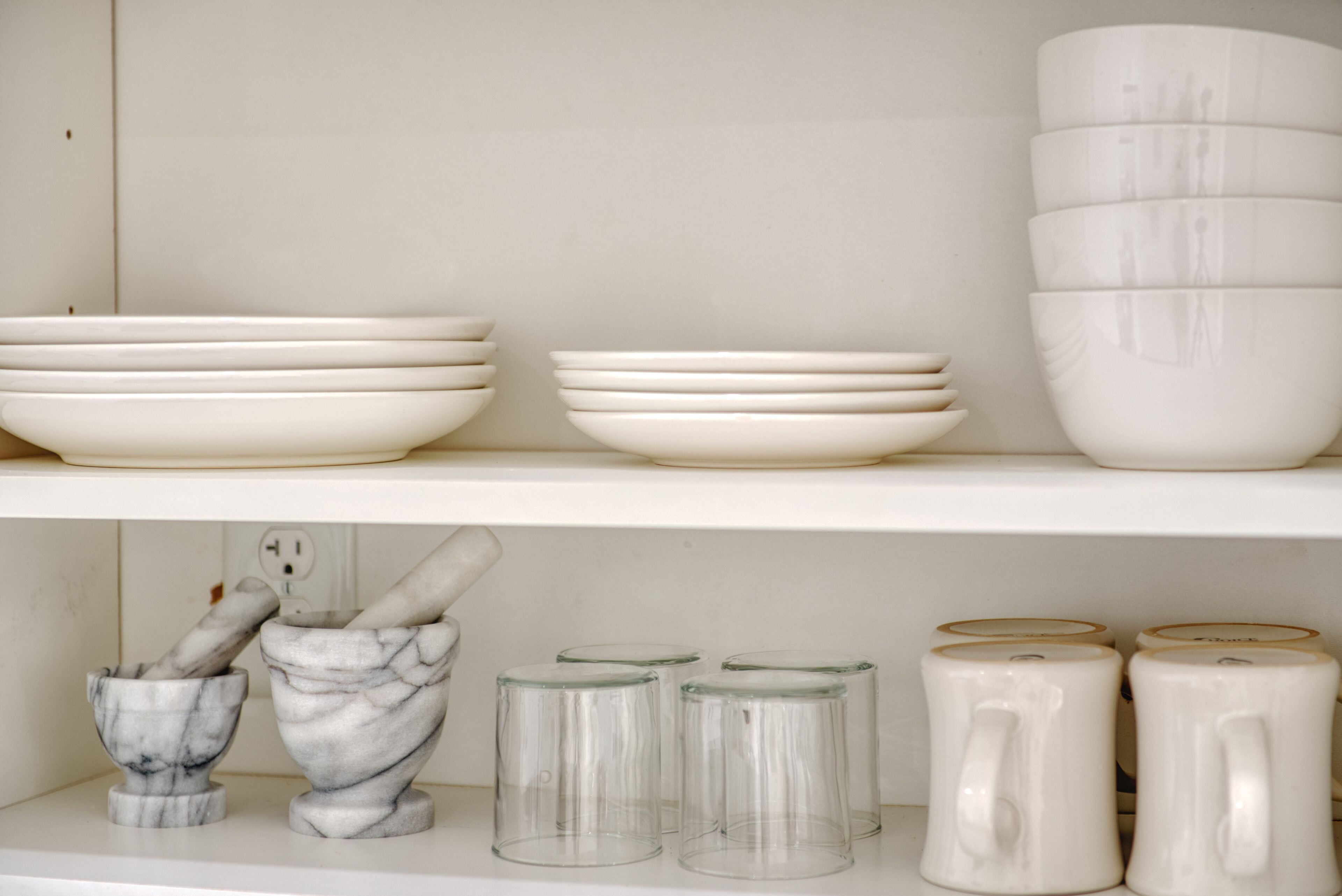 A kitchen shelf with white ceramic plates, bowls, and a marble mortar and pestle.