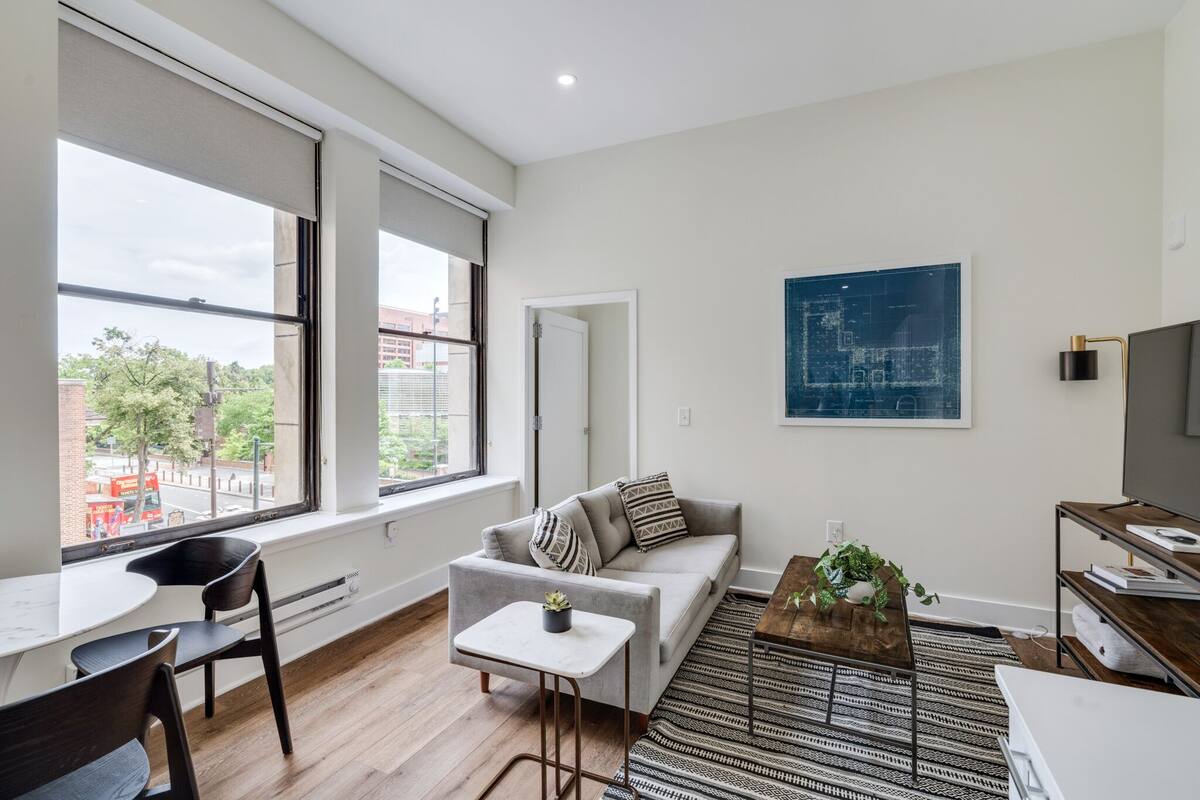Ein modernes Wohnzimmer mit Sofa, Couchtisch und Sesseln. Ein großes Fenster bietet einen Blick nach draußen, an der Wand hängt ein Gemälde und ein Fernseher steht auf einem Schrank.