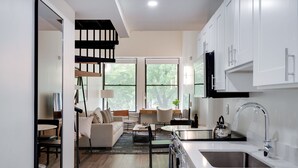 Una cocina moderna con armarios blancos, un lavamanos de acero inoxidable y una tetera negra. Hay una zona de comedor con un sofá y una mesita de centro.