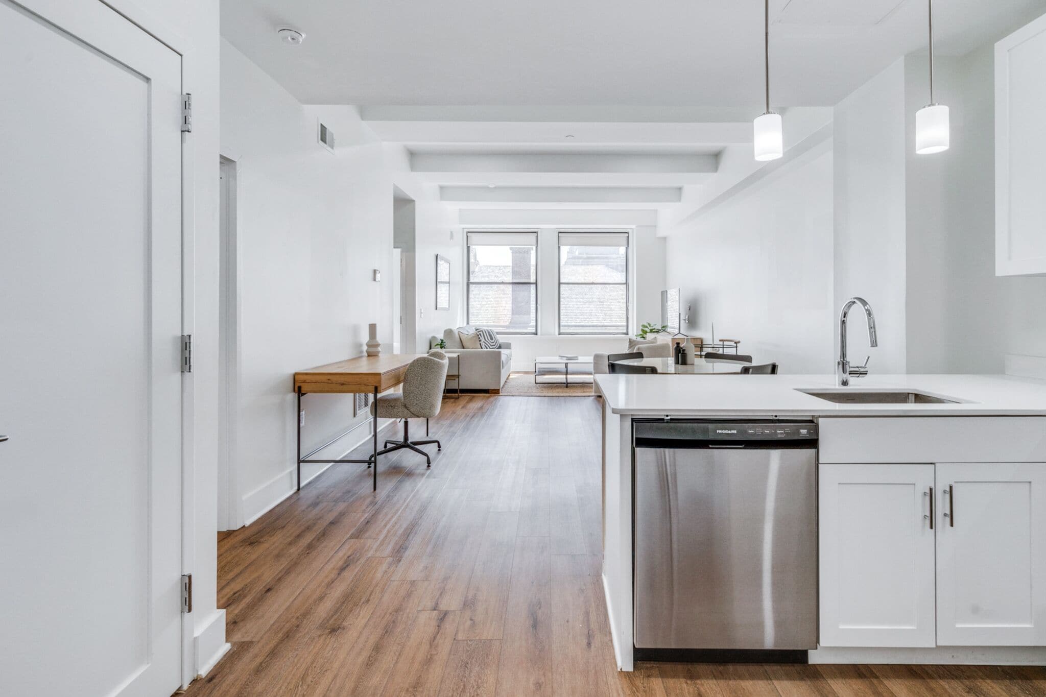 A modern kitchen with white cabinetry, a dishwasher, and a sink. There is a dining area with a table and chairs, and a large window providing natural light.
