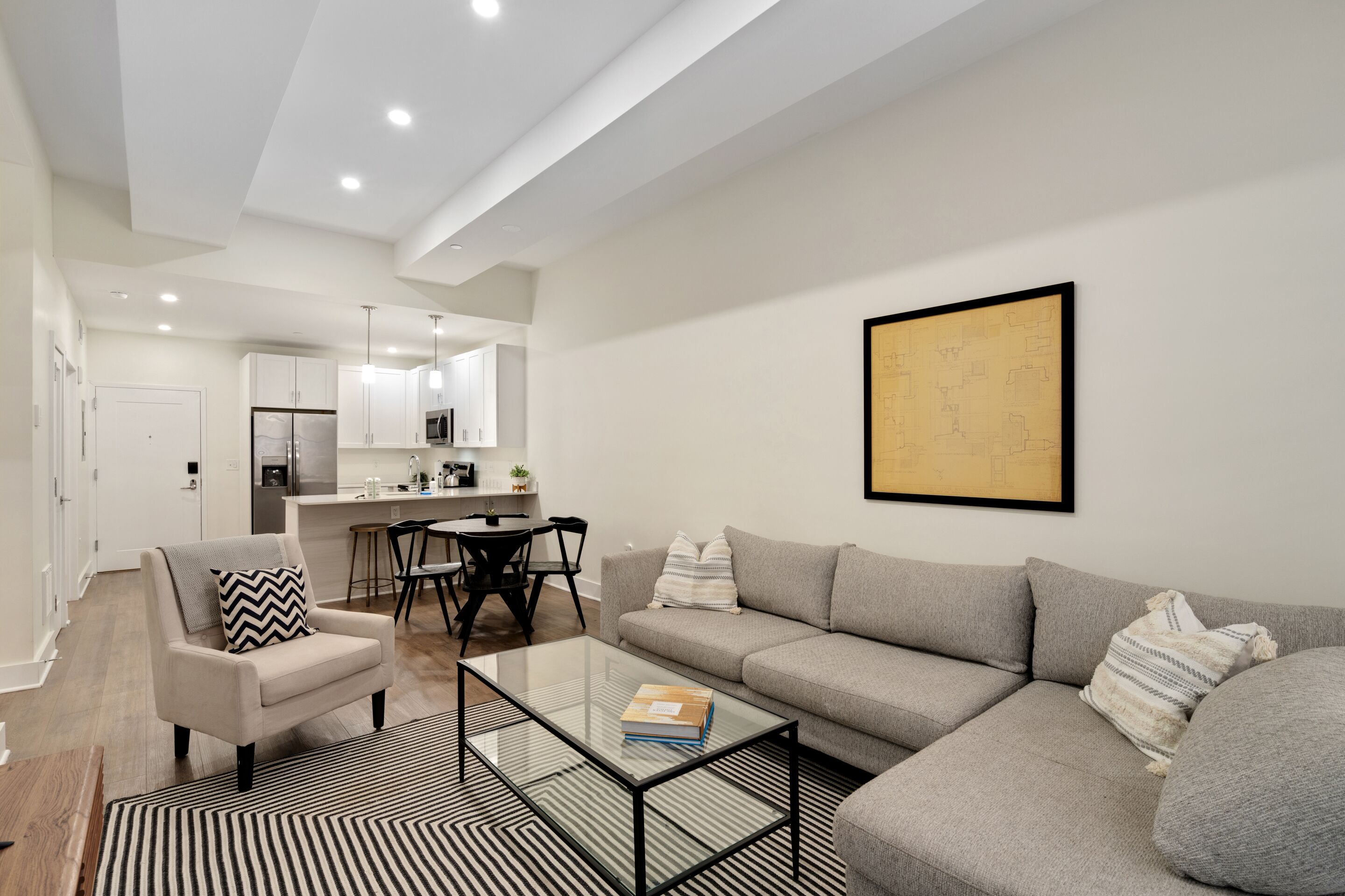 Una sala de estar moderna con sofá, sillón y mesa de centro. En el fondo, se observa un comedor con mesa y sillas.