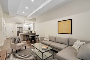 Una sala de estar moderna con sofá, sillón y mesa de centro. En el fondo, se observa un comedor con mesa y sillas.