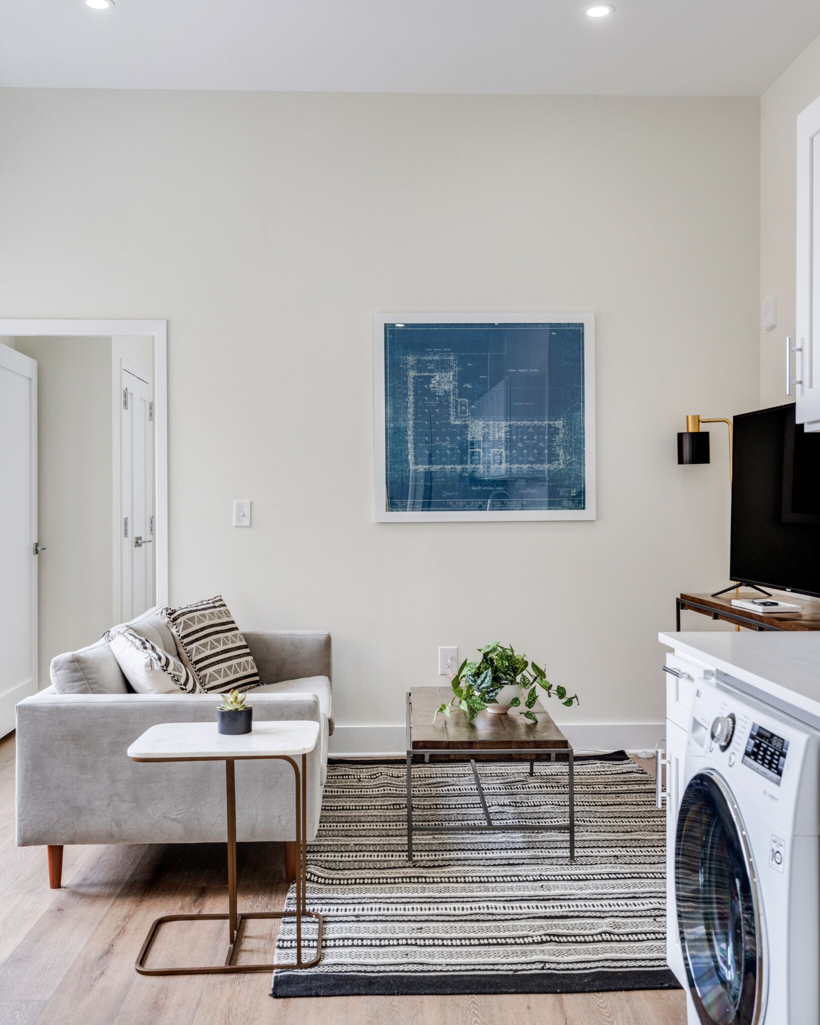 Un salon moderne avec un canapé gris, une table basse, un plan architectural encadré, un lave-linge et un téléviseur.