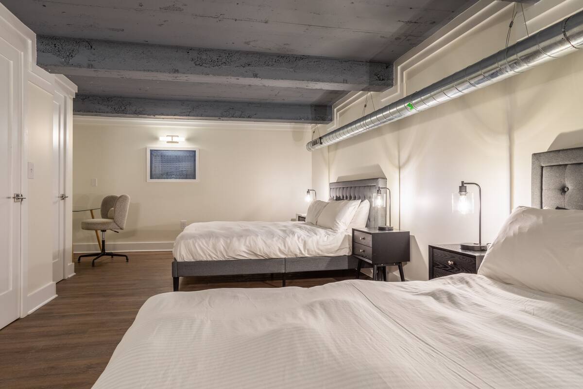 Ein Schlafzimmer mit zwei Betten, einem Schreibtisch und einem Stuhl. Die Decke hat sichtbare Holzbalken und einen Lüftungsschlitz.
