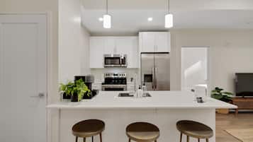 Una cocina moderna con armarios blancos, electrodomésticos de acero inoxidable, una isla central con taburetes e iluminación colgante.