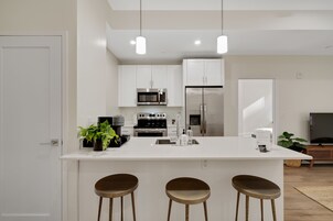 Una cocina moderna con armarios blancos, electrodomésticos de acero inoxidable, una isla central con taburetes e iluminación colgante.