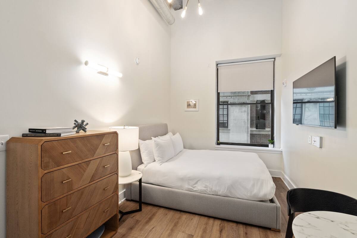 Ein modernes Schlafzimmer mit Bett, Kommode, Nachttisch, Lampe und einem an der Wand befestigten Fernseher.