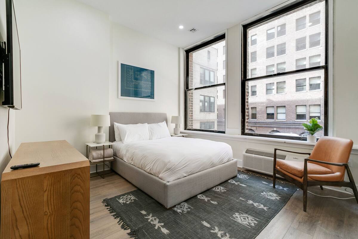 Ein modernes Schlafzimmer mit einem großen Bett, einem Holztisch, einem Stuhl und einem Fenster mit Blick auf die Stadt.