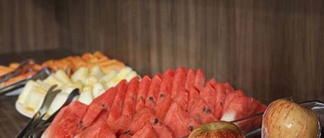 Buffet com diversos alimentos, incluindo fatias de melancia, maçãs e outras frutas.