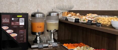 Un bufé de desayuno con una máquina de café, dispensadores de jugo y una variedad de frutas y pasteles.