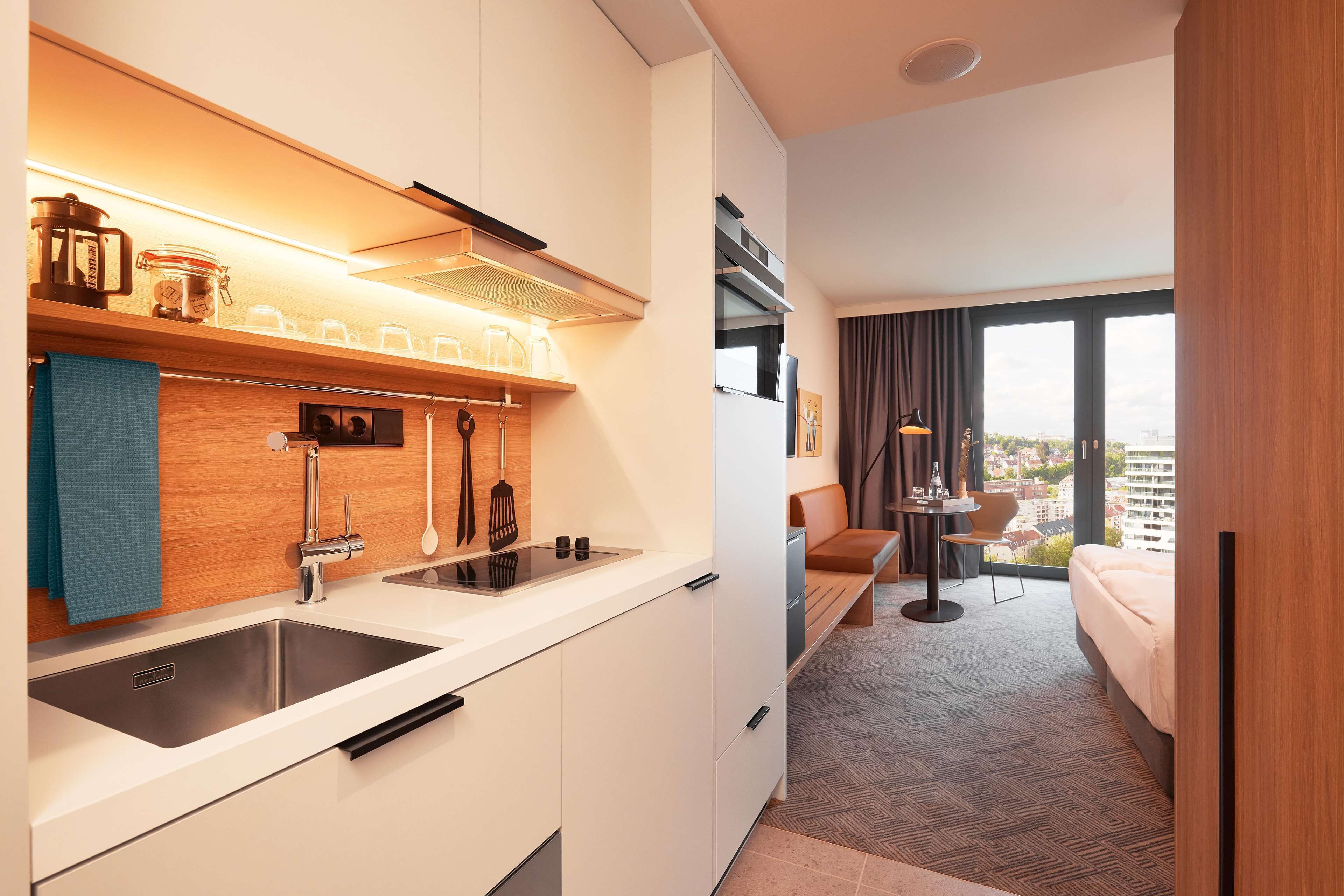 A modern hotel room with a kitchenette, a bed, a seating area, and a large window offering a city view.