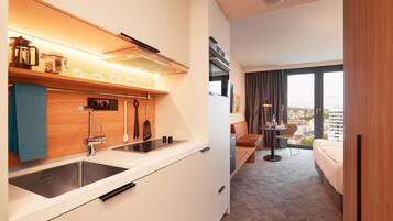 A modern hotel room with a kitchenette, a bed, a seating area, and a large window offering a city view.
