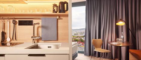 Une chambre d’hôtel moderne dotée d’un coin cuisine, d’une table ronde avec une lampe, d’une vue sur la ville et d’une grande fenêtre avec des rideaux.