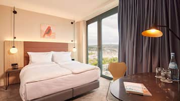 A hotel room with a bed, a table with a magazine, a bottle, and a view of the city through large windows.