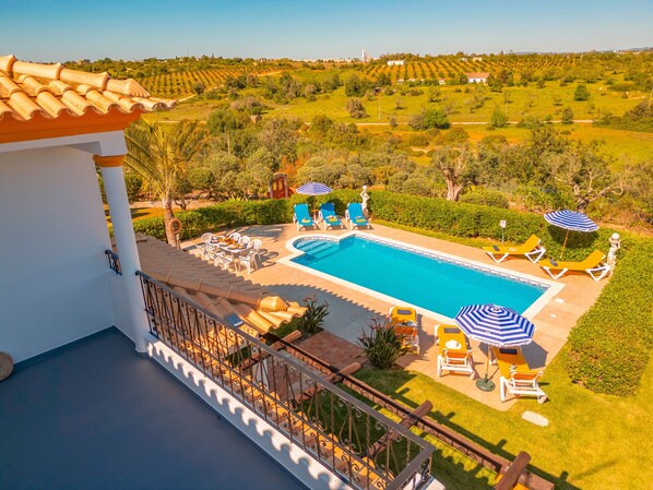 A pool area with lounge chairs and umbrellas, surrounded by greenery and a view of the countryside.