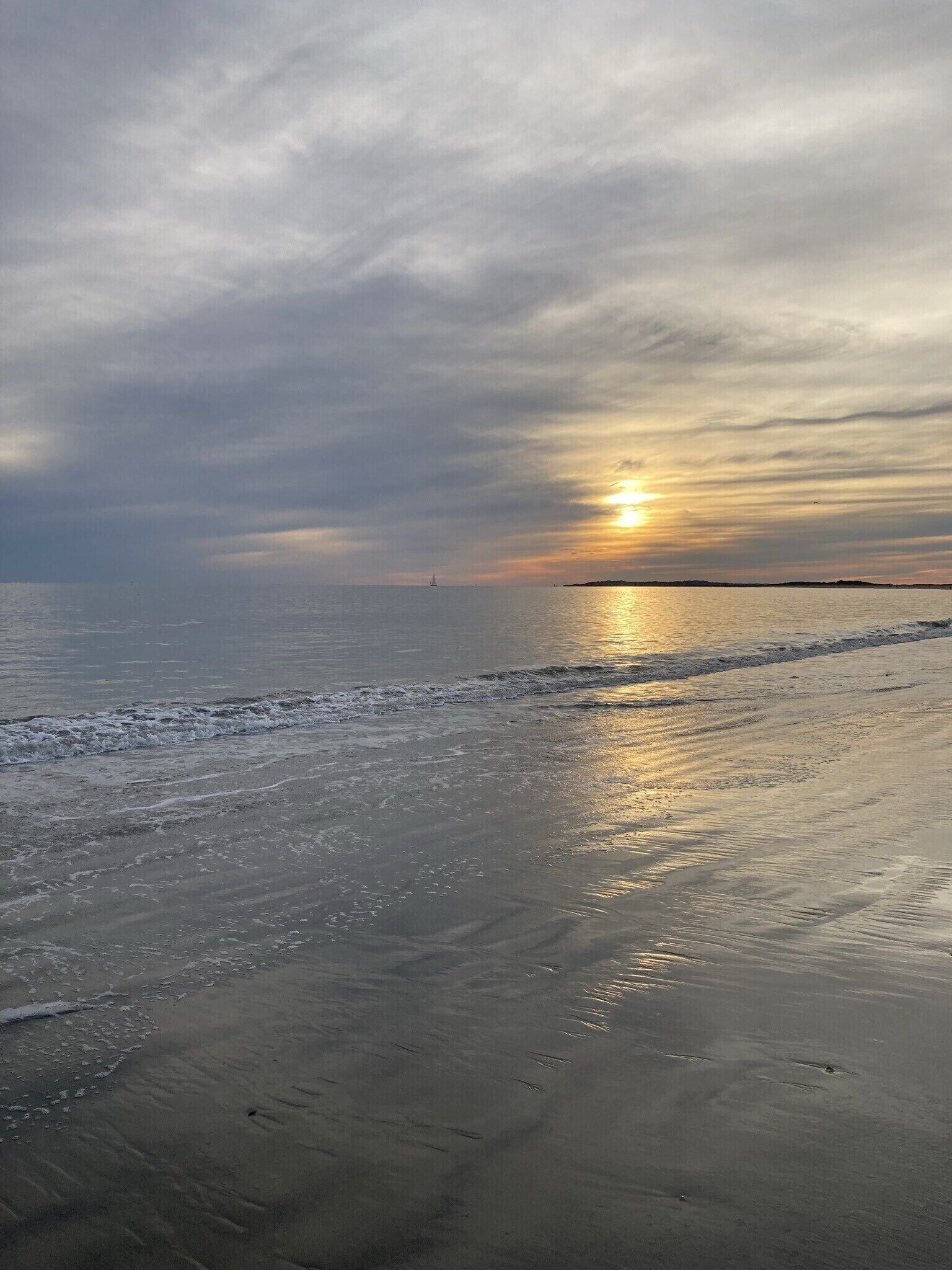 A beach with a sunset over the ocean, waves gently lapping at the shore, and a distant sailboat.