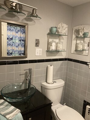 Un baño con un lavabo de vidrio, un inodoro y dos repisas con adornos.