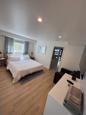 A modern hotel room with a large bed, a desk, a chair, and a door leading to another room.