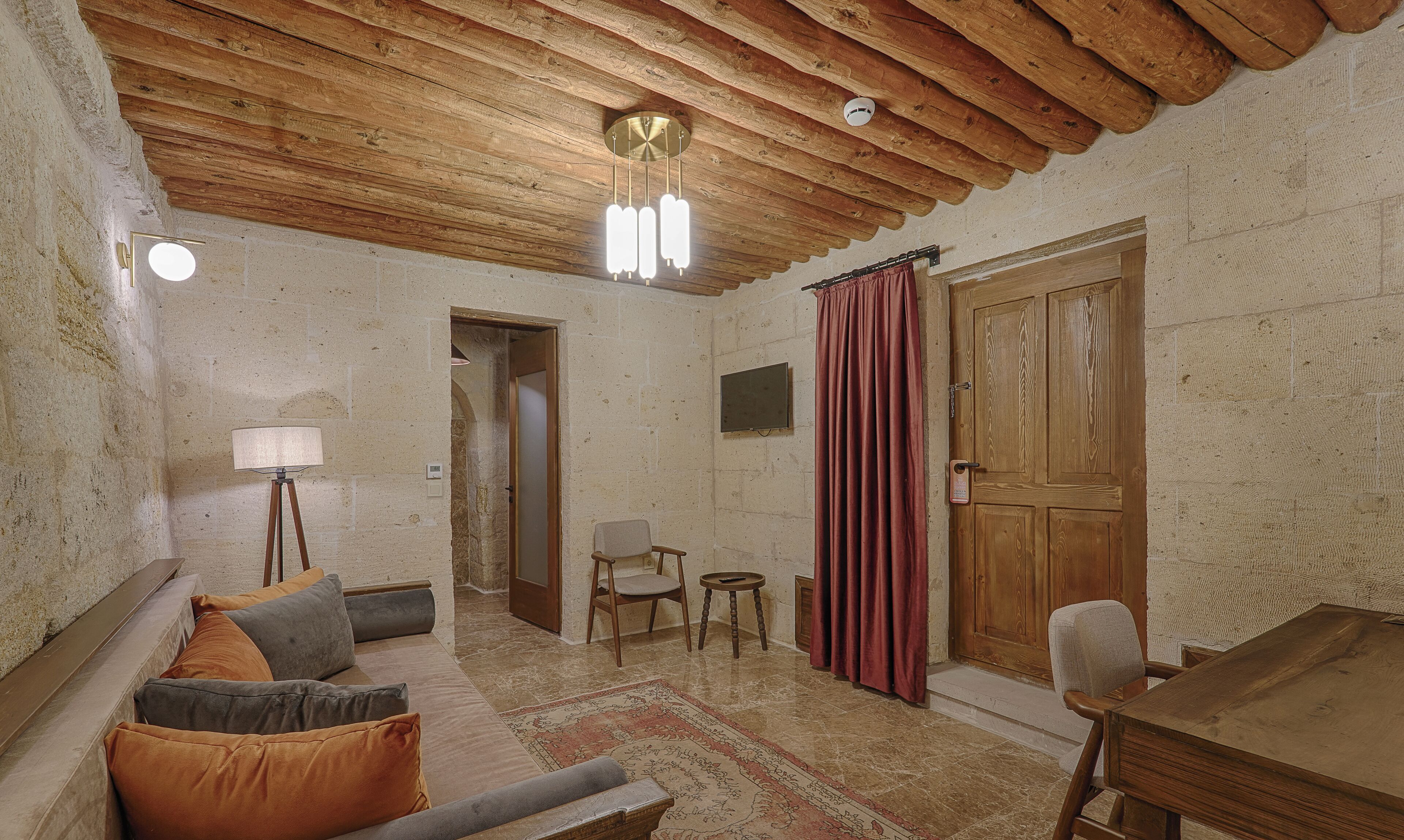 Una habitación rústica con vigas de madera a la vista, una puerta de madera, un sofá con cojines, una mesita, una silla, una lámpara de pie y un televisor.