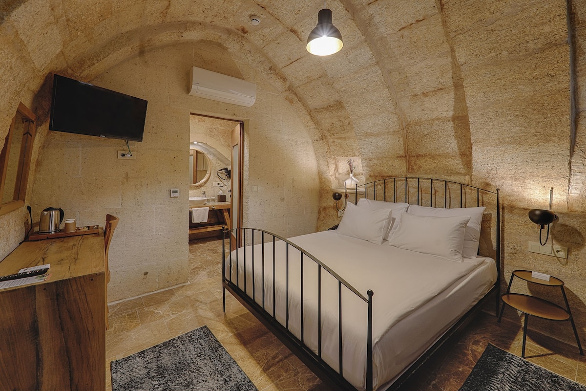 A bedroom with a bed, a desk, a chair, a TV mounted on the wall, and a lamp on the bedside table.