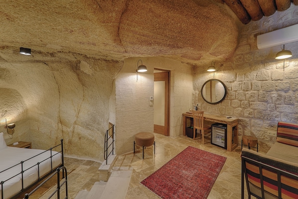 Un interior rústico de piedra con una cama, un escritorio de madera, una silla y una alfombra roja.