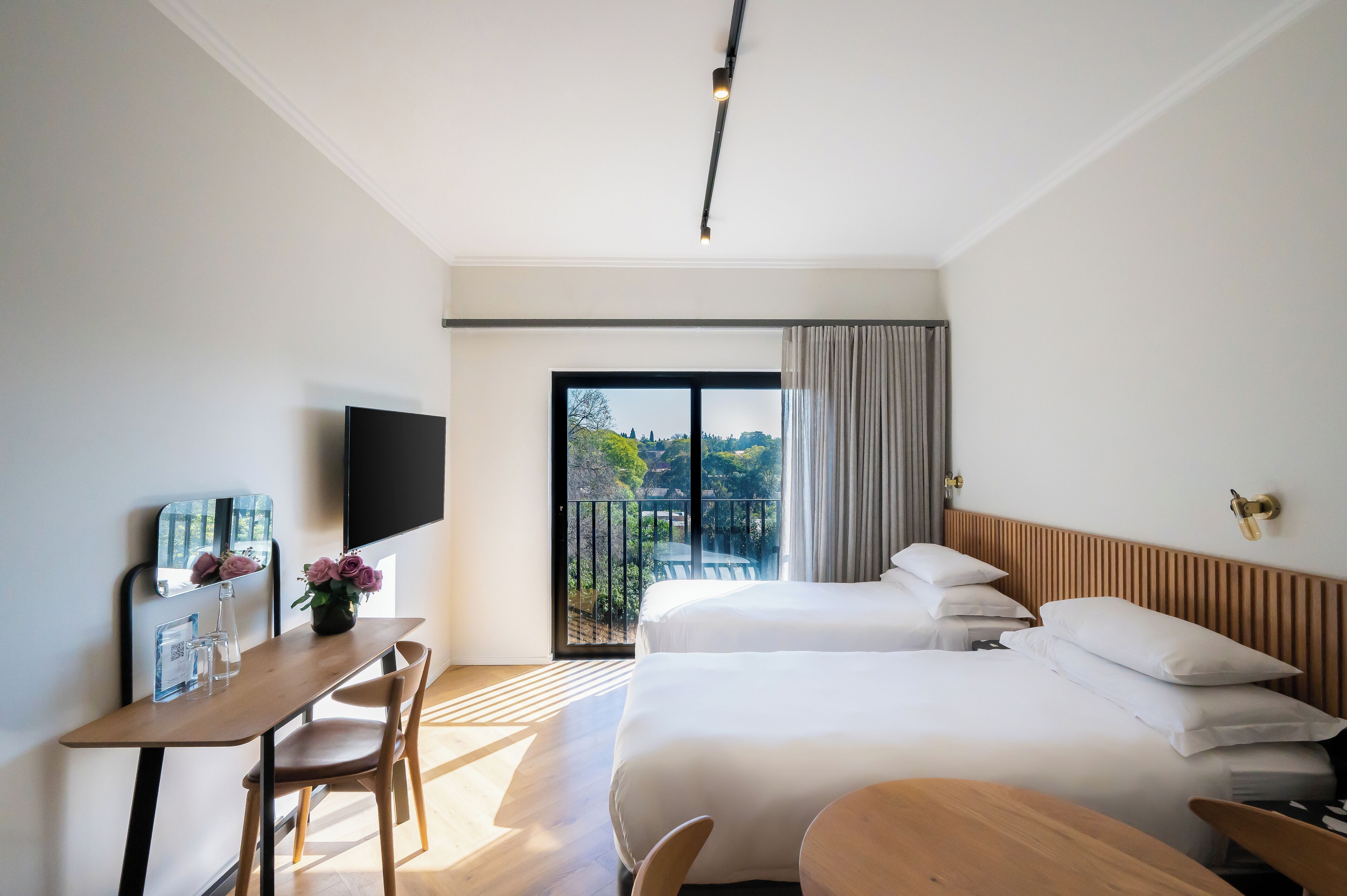 A modern hotel room with a large bed, a desk with a flat-screen TV, and a balcony with a view of greenery.
